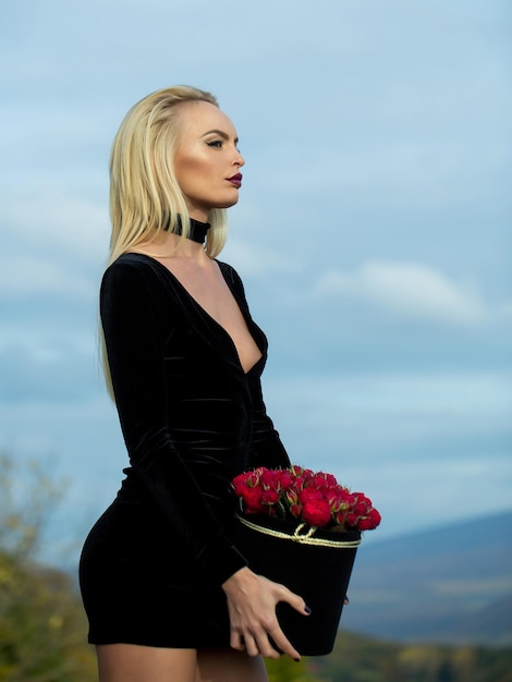 Chica muy linda o hermosa mujer rubia en sexy vestido negro con flores rosas rojas en caja al aire libre sobre fondo natural con cielo azul nublado