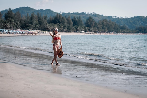 Chica muy rubia vestida con un sombrero de paja y bikini rojo en un estado de ánimo feliz en la playa. Phuket. Tailandia Vacaciones de verano. Fondo marino