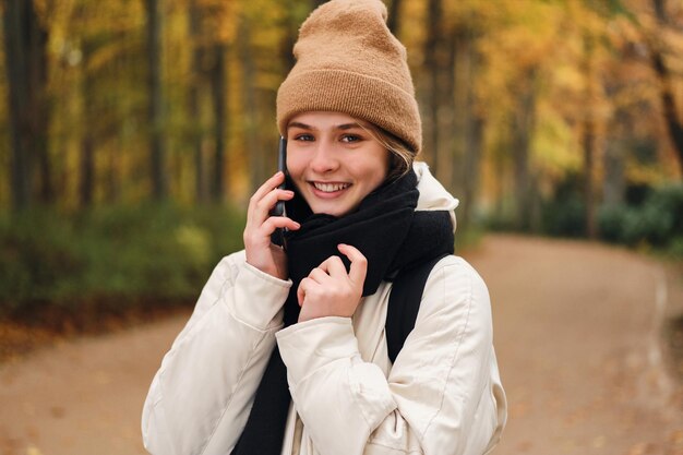 Chica muy sonriente hablando por teléfono celular mirando alegremente a la cámara en el hermoso parque de otoño