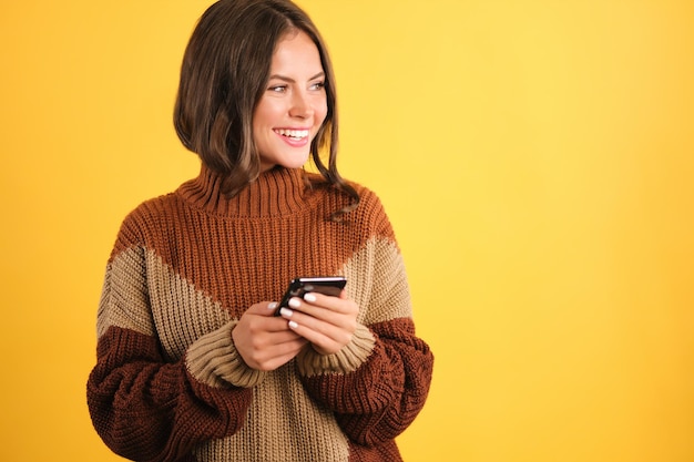 Chica muy sonriente en suéter acogedor con teléfono celular mirando alegremente a un lado sobre fondo amarillo