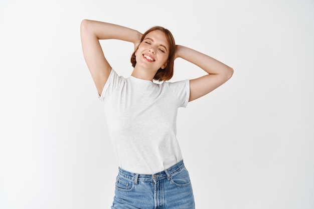 Chica natural despreocupada que se siente descansada y relajada, sosteniendo las manos detrás de la cabeza, sonriendo y estirándose con los ojos cerrados, de pie contra la pared blanca