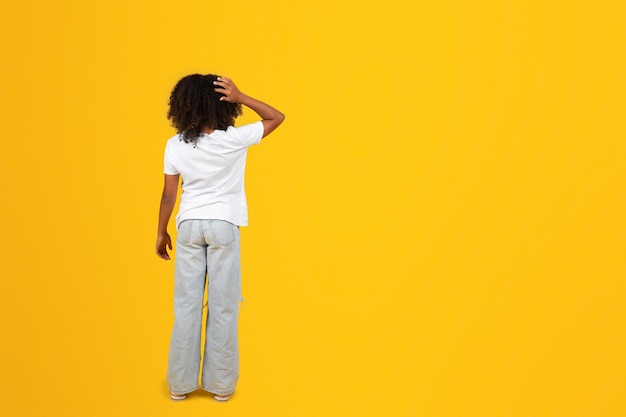 Chica negra adolescente pensativa con camiseta blanca se rasca la cabeza con la mano aislada en el fondo amarillo