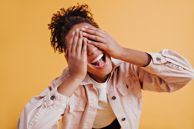 Chica negra emocionada con cola de caballo que cubre los ojos con las manos Foto de estudio de una mujer africana alegre y sonriente con chaqueta