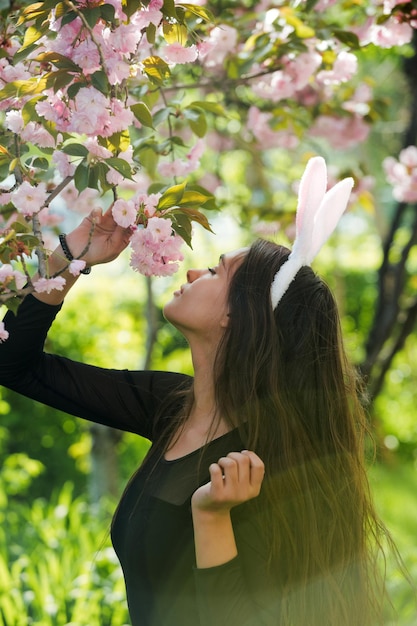 Chica con orejas de conejo rosadas que huelen las flores de sakura florecientes en primavera parque primavera de pascua