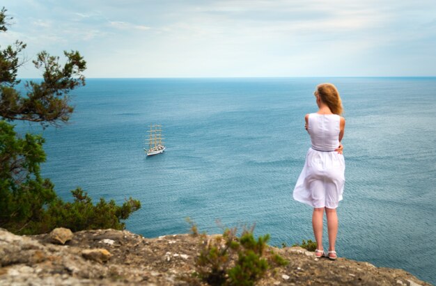 Chica en la orilla del mar mira a lo lejos en un velero en el horizonte, el concepto de expectativa, esperanza, amor, anhelo, tristeza, vista posterior.