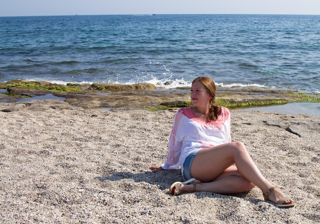 Chica en pantalones cortos y blusa blanca en la playa junto al mar .. Mujer pelirroja en una playa de arena. Unas vacaciones en el sur