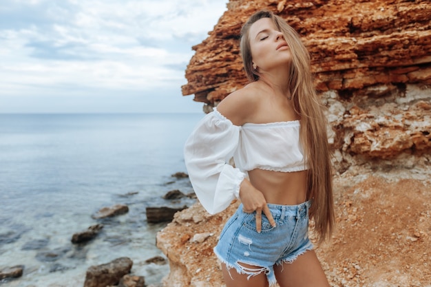 Una chica en pantalones cortos de mezclilla y una blusa blanca transparente en la playa.