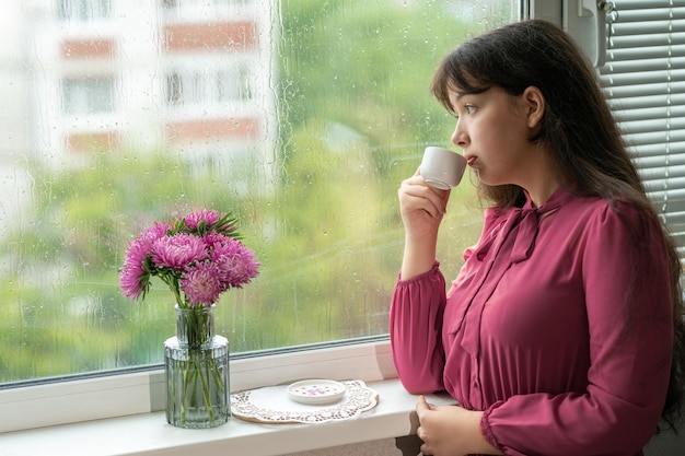 La chica parada al lado de una ventana lluviosa y bebe un café.