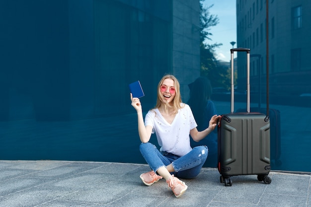 Chica con pasaporte y equipaje en gafas de color rosa sobre un fondo azul.