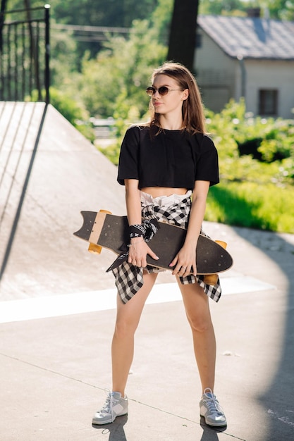 Chica patinadora adolescente sosteniendo patineta y de pie en el parque de patinadores