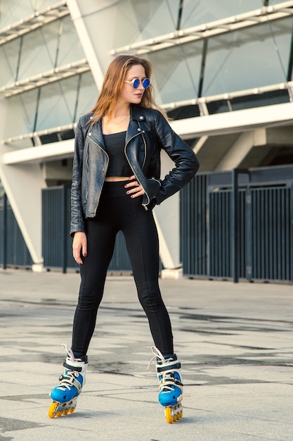 Chica en patines de pie en el fondo del edificio. Chica joven en forma de mujer con gafas de sol azules, jeans y chaqueta en patines montando al aire libre después de la lluvia.