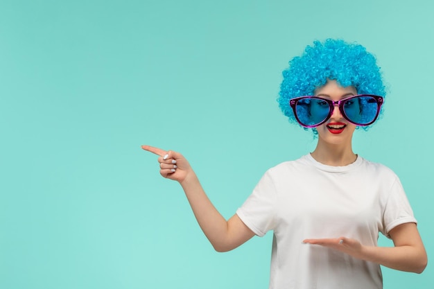 Chica de payaso del día de los inocentes dirigiendo el dedo a la izquierda grandes gafas de sol azules traje divertido cabello azul