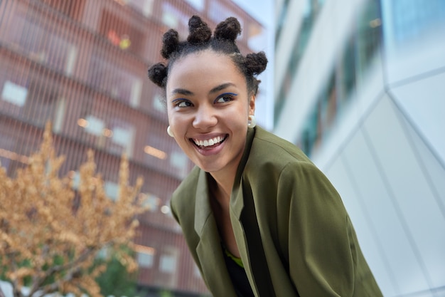 Chica con peinado de moño sonríe positivamente muestra dientes blancos uniformes vestidos con chaqueta verde concentrada poses de distancia contra el fondo borroso se divierte