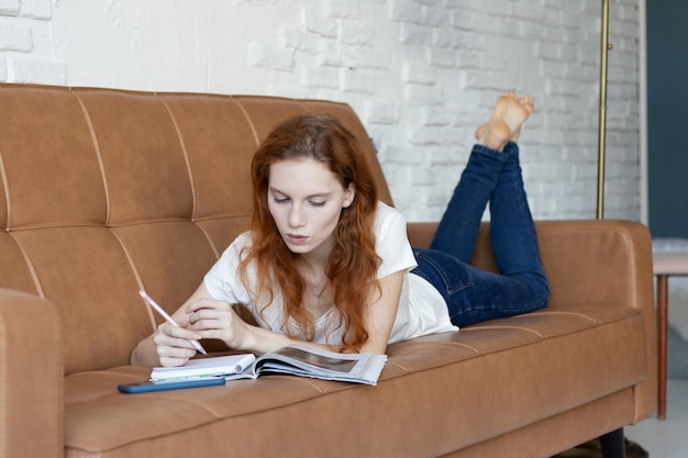 Chica pelirroja se acuesta en el sofá y lee una revista en casa