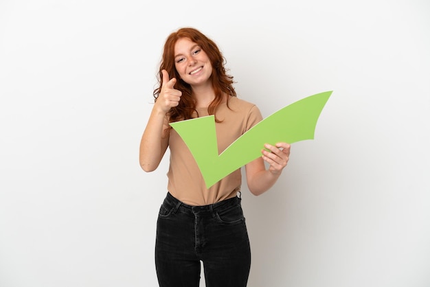 Foto chica pelirroja adolescente aislada sobre fondo blanco sosteniendo un icono de verificación y apuntando hacia el frente