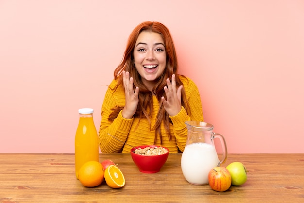 Chica pelirroja adolescente desayunando en una mesa con expresión facial sorpresa