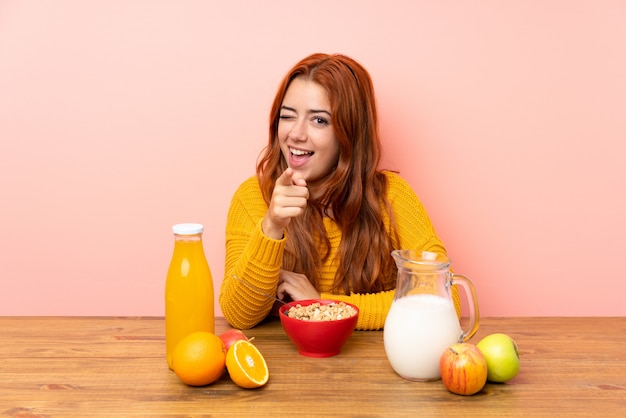 Chica pelirroja adolescente desayunando en una mesa te señala con el dedo