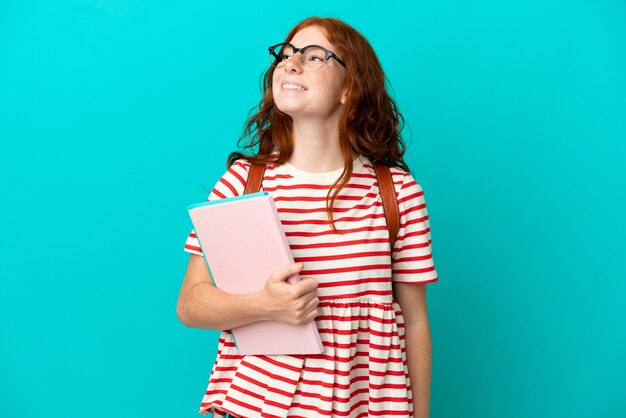 Chica pelirroja adolescente estudiante aislada sobre fondo azul pensando en una idea mientras mira hacia arriba