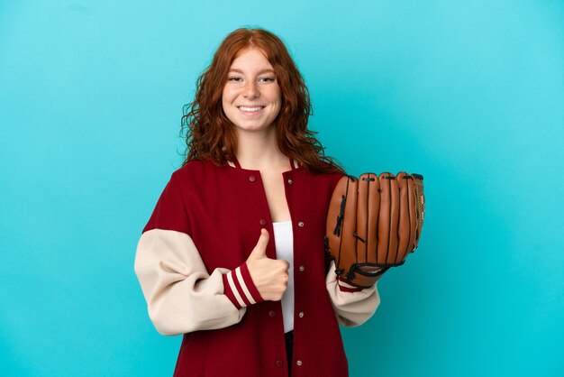 Chica pelirroja adolescente con guante de béisbol aislado sobre fondo azul dando un pulgar hacia arriba gesto