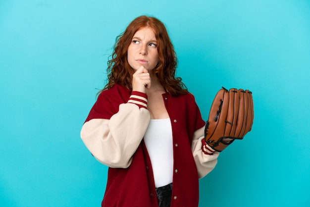 Chica pelirroja adolescente con guante de béisbol aislado sobre fondo azul teniendo dudas y pensando