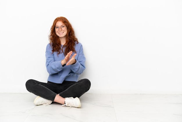 Chica pelirroja adolescente sentada en el suelo aislado sobre fondo blanco aplaudiendo después de la presentación en una conferencia