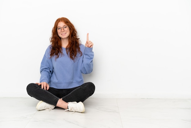 Chica pelirroja adolescente sentada en el suelo aislado sobre fondo blanco mostrando y levantando un dedo en señal de lo mejor