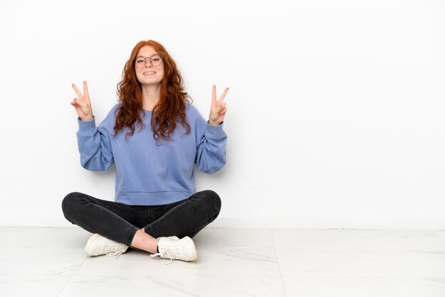 Chica pelirroja adolescente sentada en el suelo aislado sobre fondo blanco mostrando el signo de la victoria con ambas manos