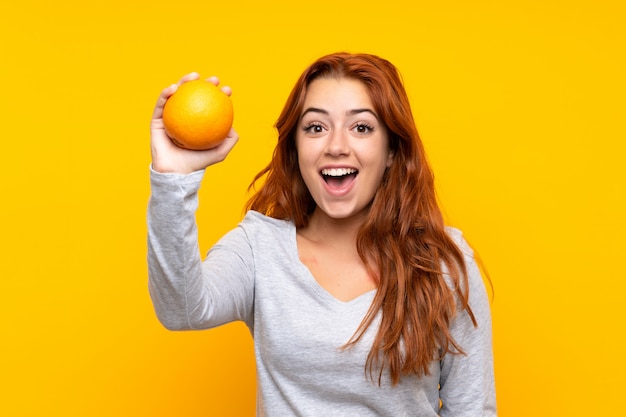 Foto chica pelirroja adolescente sosteniendo una naranja sobre amarillo aislado