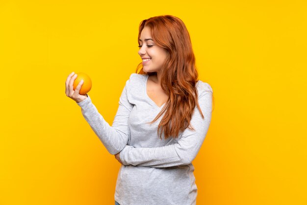 Chica pelirroja adolescente sosteniendo una naranja sobre fondo amarillo aislado