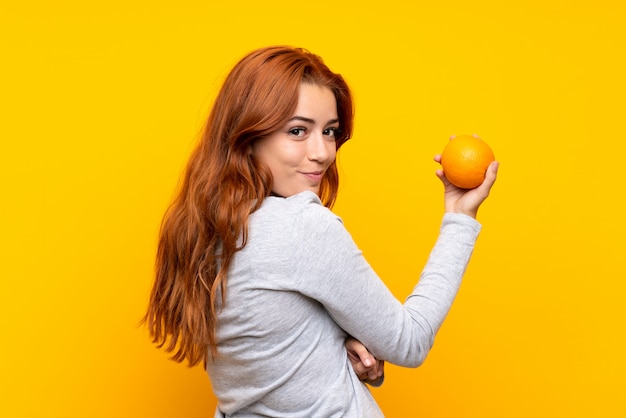 Chica pelirroja adolescente sosteniendo una naranja sobre pared amarilla aislada