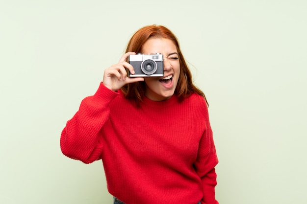 Chica pelirroja adolescente con suéter sobre pared verde aislado sosteniendo una cámara