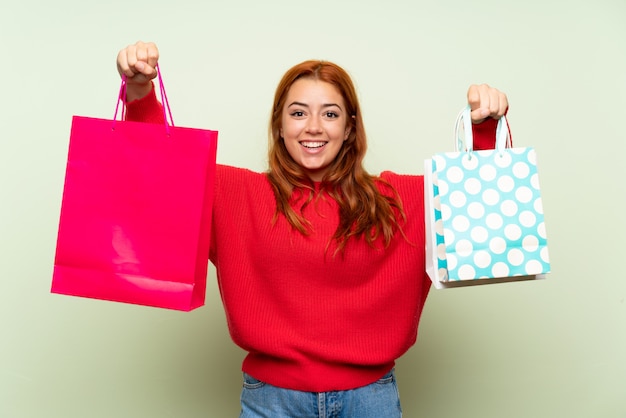 Chica pelirroja adolescente con suéter sobre pared verde aislado sosteniendo muchas bolsas de compras