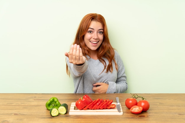 Chica pelirroja adolescente con verduras en una mesa invitando a venir