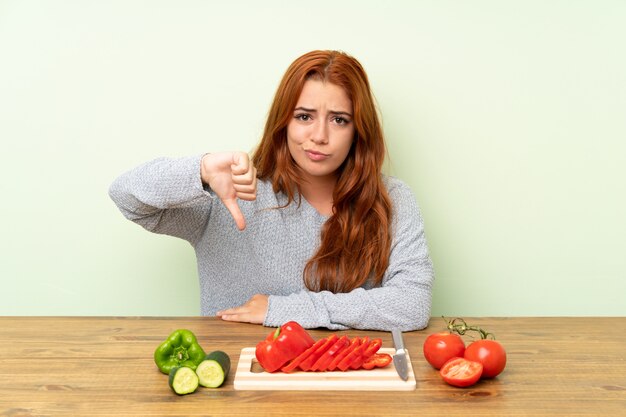 Chica pelirroja adolescente con verduras en una mesa mostrando el pulgar hacia abajo signo