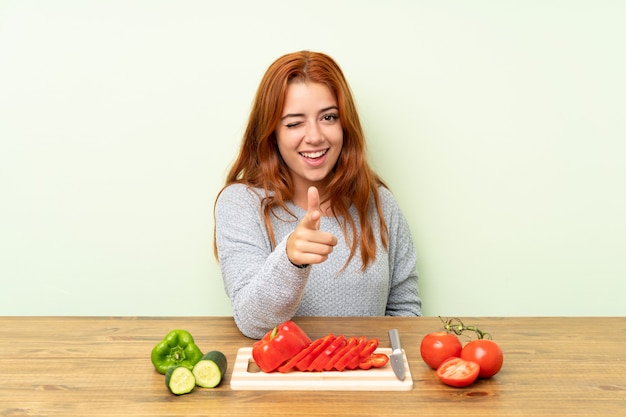 Chica pelirroja adolescente con verduras en una mesa te señala con el dedo