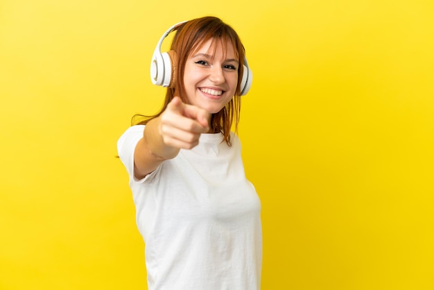 Chica pelirroja aislada de fondo amarillo escuchando música y apuntando al frente