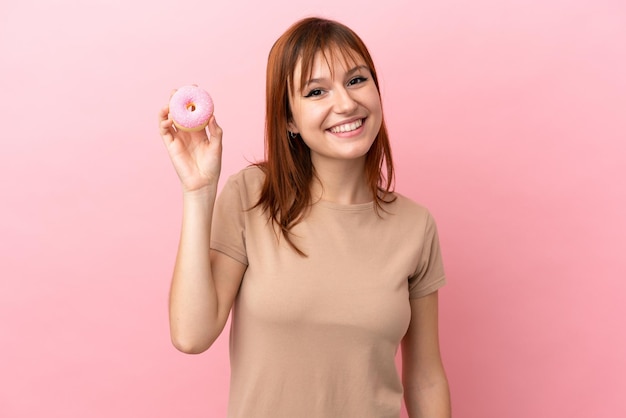 Chica pelirroja aislada sobre fondo rosa sosteniendo un donut y feliz