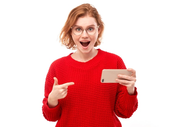 Chica pelirroja alegre sorprendida señala con el dedo a la pantalla en blanco del teléfono inteligente con gafas y ropa roja aislada sobre fondo blanco de estudio