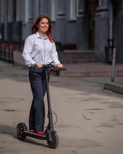 Una chica pelirroja con una camisa blanca conduce un scooter eléctrico a lo largo de la pared. Una mujer de negocios con traje de pantalón y tacones rojos recorre la ciudad en un moderno código de vestimenta en la oficina.