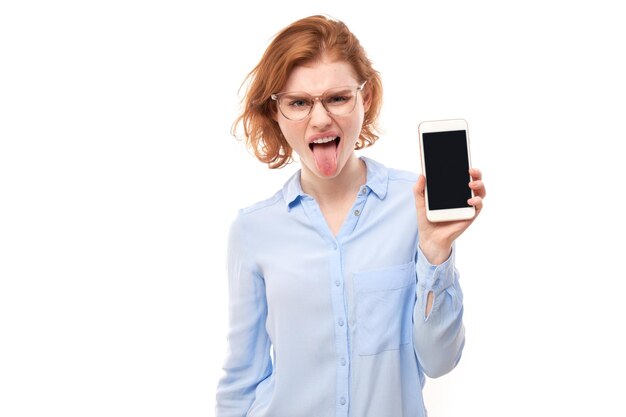 Chica pelirroja con cara de disgusto demuestra la pantalla en blanco del teléfono inteligente y muestra la lengua en gafas y camisa de negocios aislada sobre fondo blanco de estudio