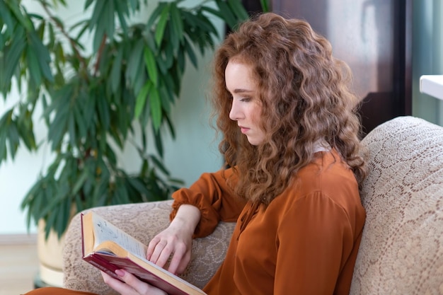 Chica pelirroja en casa leyendo un libro en un sofá acogedor