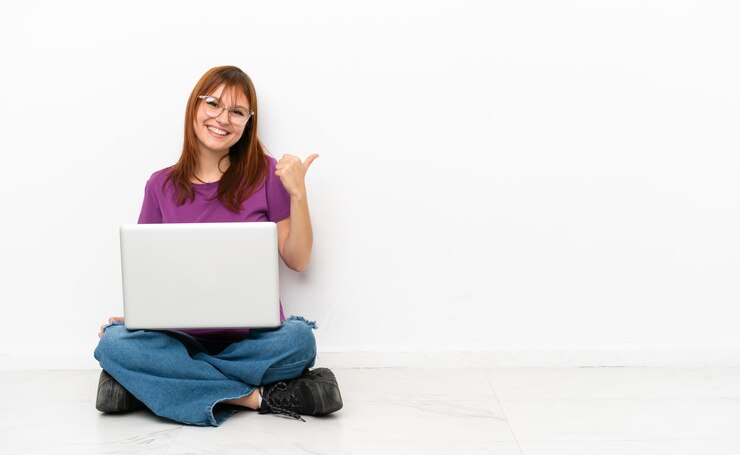 Foto chica pelirroja con una computadora portátil sentada en el suelo apuntando hacia un lado para presentar un producto