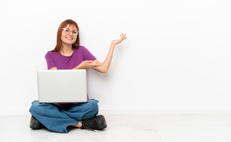 Foto chica pelirroja con una computadora portátil sentada en el suelo extendiendo las manos a un lado para invitar a venir