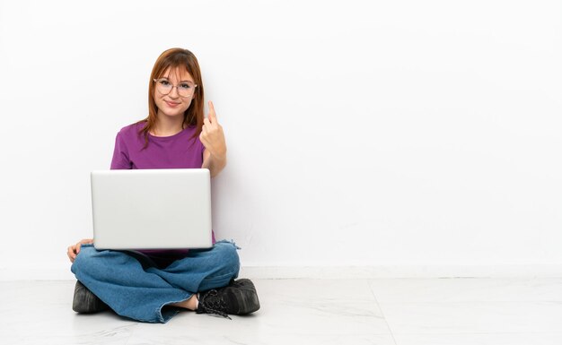 Chica pelirroja con una computadora portátil sentada en el suelo haciendo el gesto de venir