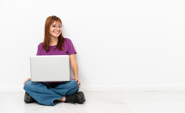 Chica pelirroja con una computadora portátil sentada en el suelo mirando hacia un lado y sonriendo