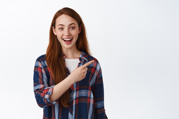 Chica pelirroja emocionada con el pelo lacio y una sonrisa blanca, apuntando con el dedo directamente al logotipo, mostrando un anuncio en un espacio vacío, de pie contra un fondo blanco