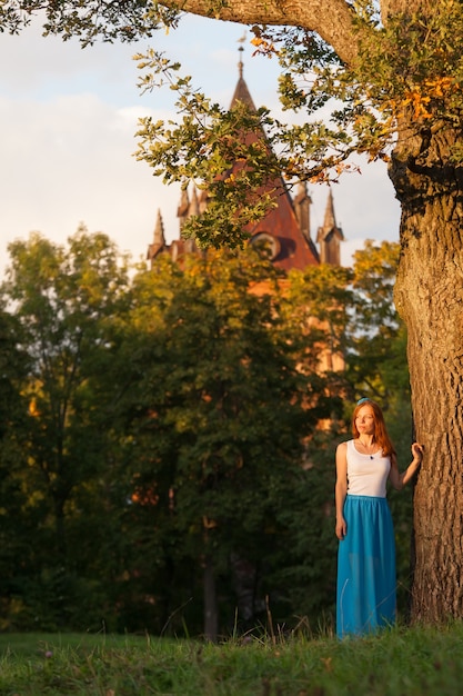 Chica pelirroja se encuentra cerca de roble viejo en el parque antes de la torre gótica