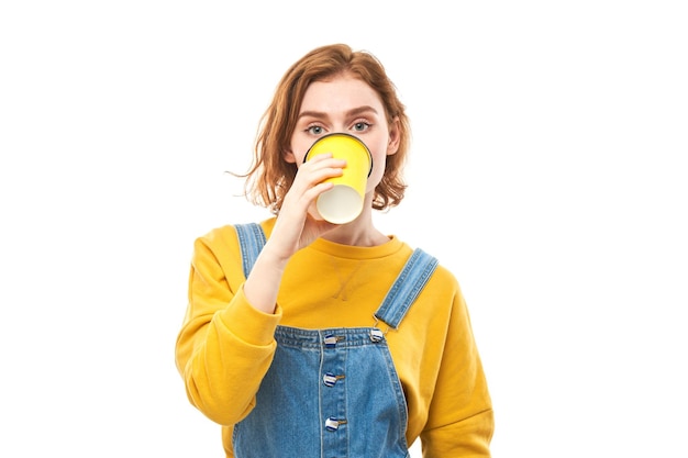 Chica pelirroja enérgica sosteniendo y bebiendo una taza amarilla de café para llevar sonriendo y disfrutando de una bebida aislada de fondo blanco