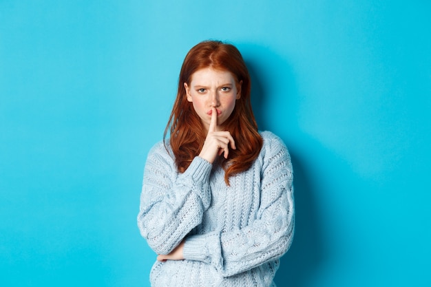 Chica pelirroja enojada que se está callando, muestra un gesto tabú, prohíbe hablar, de pie sobre un fondo azul en un suéter