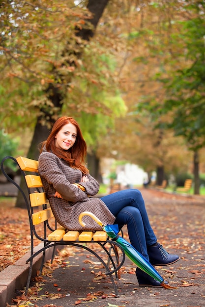 Chica pelirroja de estilo sentada en un banco en el parque de otoño.